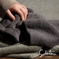 兩條深灰色與淺灰色質地蓬鬆柔軟的浴巾，疊放於桌面，一隻手正觸碰其中一條浴巾。