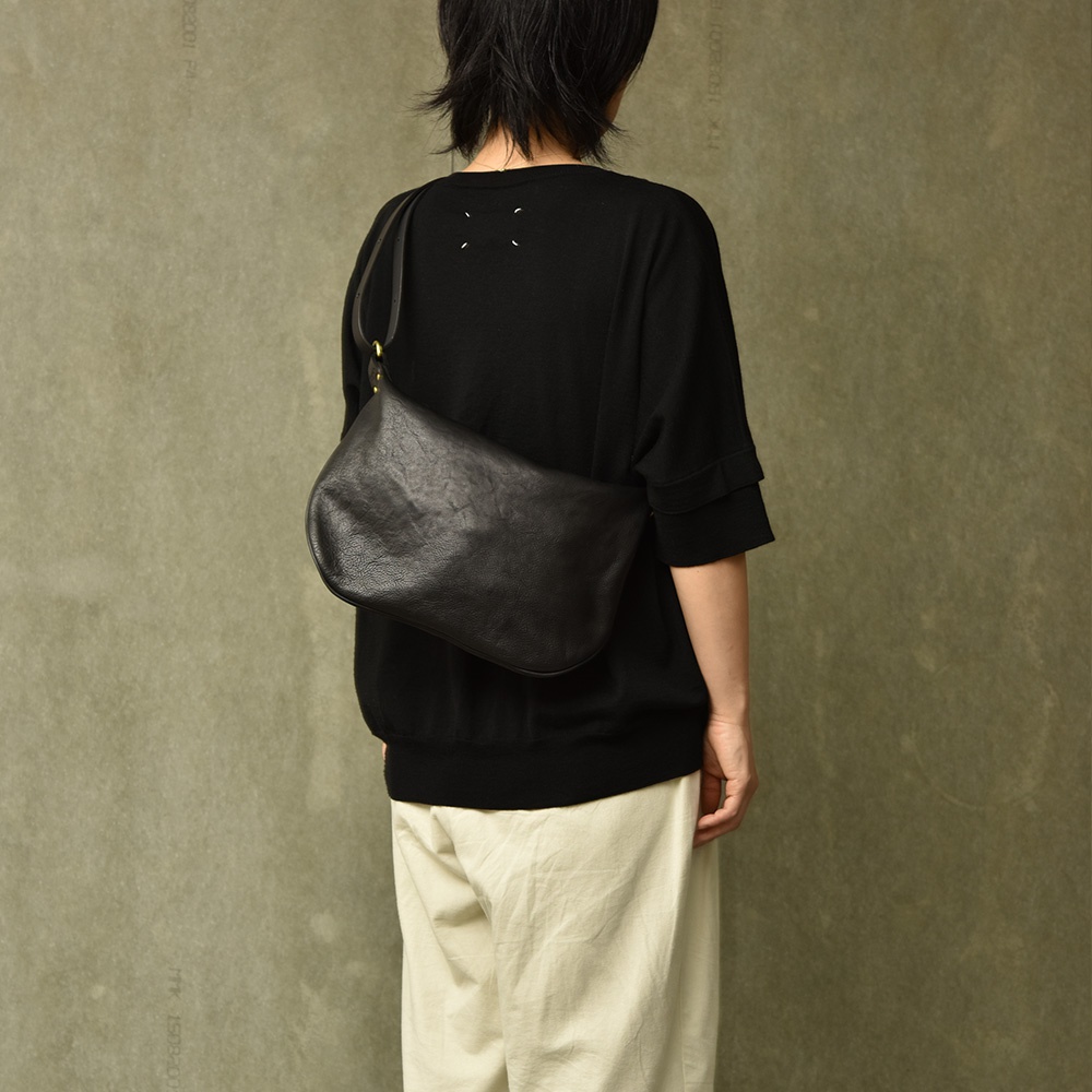 黑色皮革單肩半月包，有金色金屬扣環，由一位穿著黑色上衣和米色褲子的人背著，背景為灰色紋理牆。