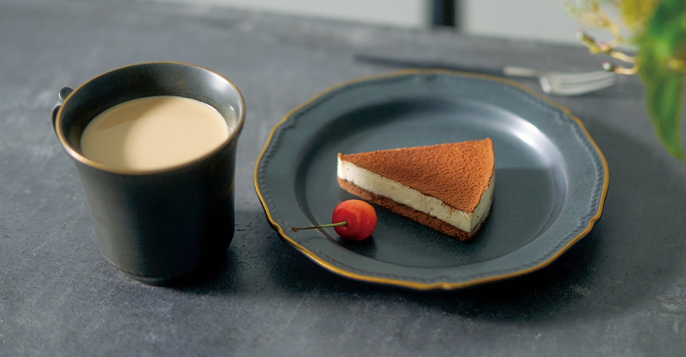 深色馬克杯裝有奶茶，旁邊有一個裝有提拉米蘇蛋糕和櫻桃的淺盤。