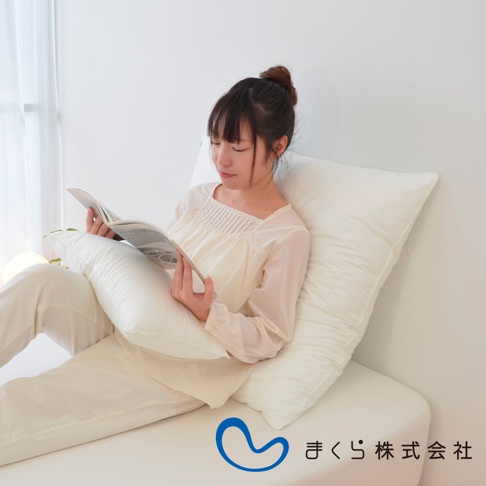 一名女子斜倚在床上，手捧一本書閱讀，身旁擺放著兩個白色方枕，其中一個枕頭放在她身前，支撐著她的手臂。