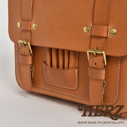 棕褐色皮革製作的學生包，有金屬扣環和鉚釘裝飾，正面設有插槽式收納袋。