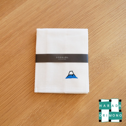 一條白色八重紗精繡擦拭巾，帶有深色紙帶，上面印有日文和英文「KAYA KUI FUKIN」，下方繡有藍色富士山圖案。