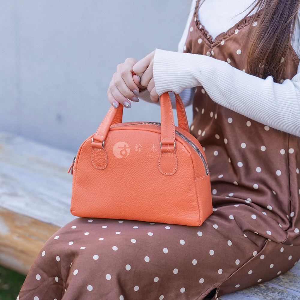 一個橘色皮革製成的半月形迷你手提包，有雙提把與拉鍊開口，包身呈現自然紋理，由一位女士持握。