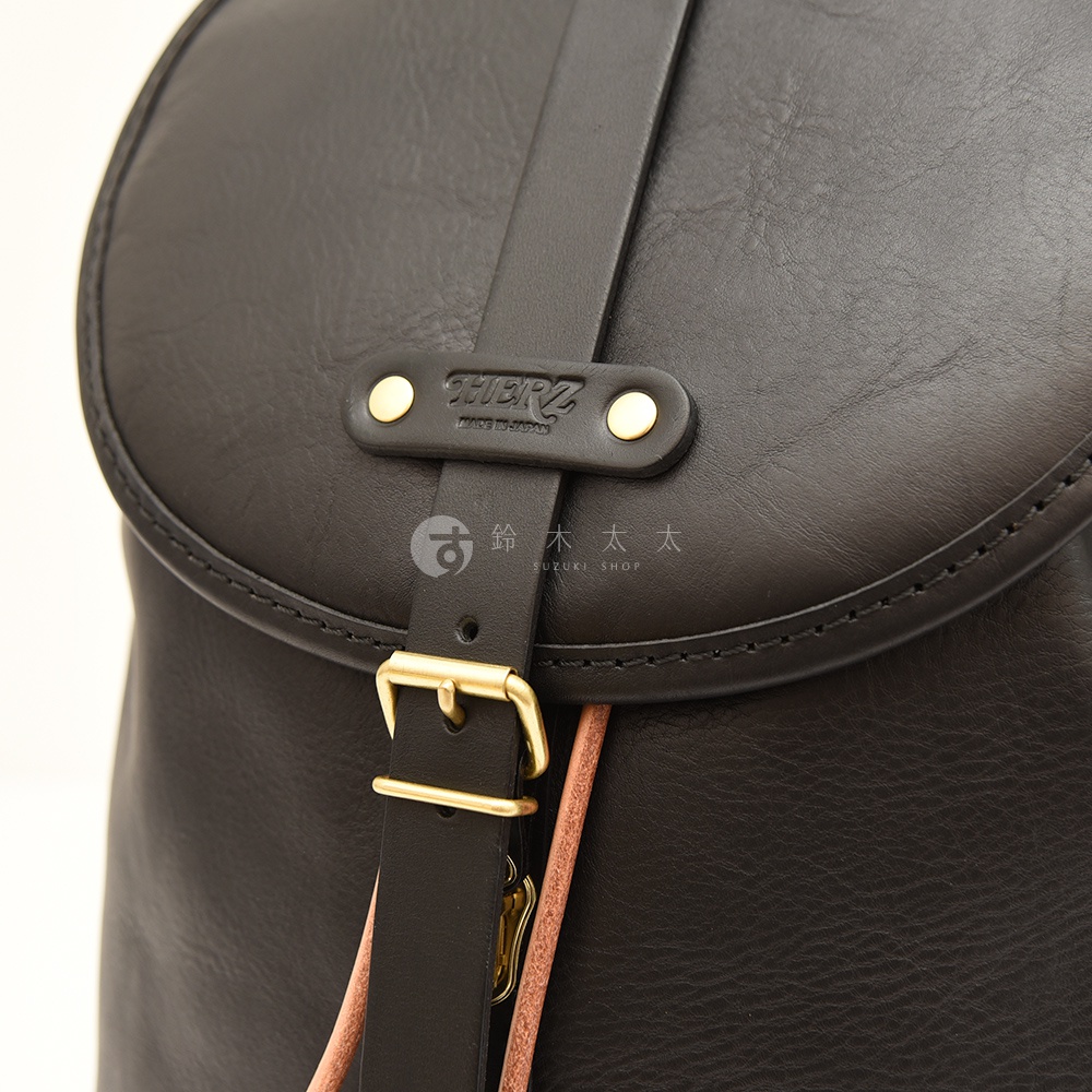 黑色皮革單扣束口後背包，特寫局部，可見品牌標誌「HERZ」及金色扣環，材質呈現自然紋理。