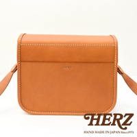 日本HERZ職人手作皮革經典雙扣郵差包，焦糖色牛皮材質，銀色雙扣設計，正面壓印HERZ品牌標誌及MADE IN JAPAN字樣，附有可調式背帶，整體呈現簡約復古風格。