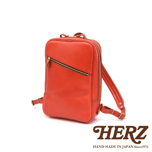 橘紅色皮革長方型後背包，具有一個斜向拉鍊前袋，兩個側邊拉鍊，以及可調節的皮革肩帶。