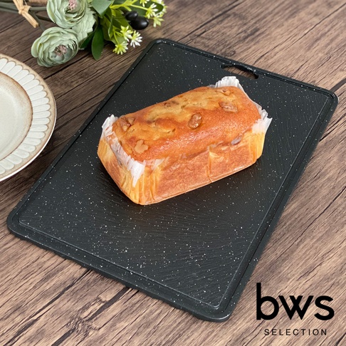 一個深灰色、帶有白色斑點的長方形食物快速冷卻盤，盤上放著一個用烘焙紙包裹的烤製麵包，麵包表面有核桃碎。