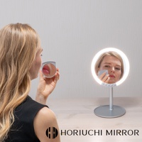 銀色金屬底座立式LED化妝鏡，圓形鏡面環繞著白色LED燈，鏡中映照著一位女士正在補妝。鏡前有一手持式迷你放大鏡，鏡頭聚焦於嘴唇。