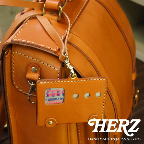 一個橙色皮革製的側背包，上面掛著一個同樣材質的卡套，卡套內有一個類似交通票券的貼紙，右下角印有 HERZ 和 HAND MADE IN JAPAN Since1973 的白色文字