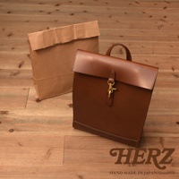 棕色皮革製成的紙袋風後背包，具有翻蓋式設計和黃銅扣環，旁邊有一個類似的棕色紙袋。