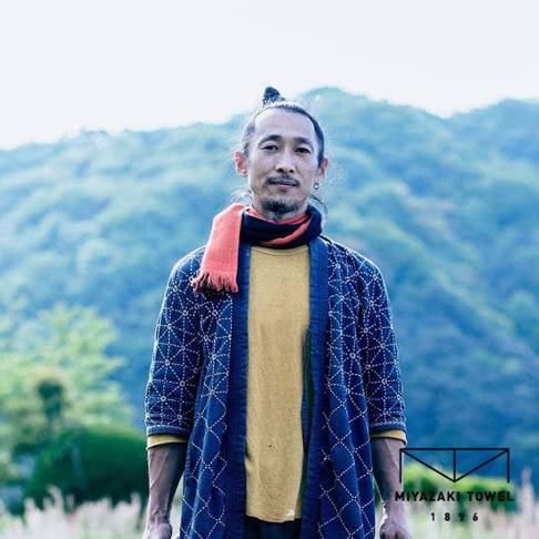 今治匹馬棉雙色圍巾，一端為橘紅色，另一端為海軍藍，圍繞在身穿黃色上衣和藍色織紋外套的男子頸部。