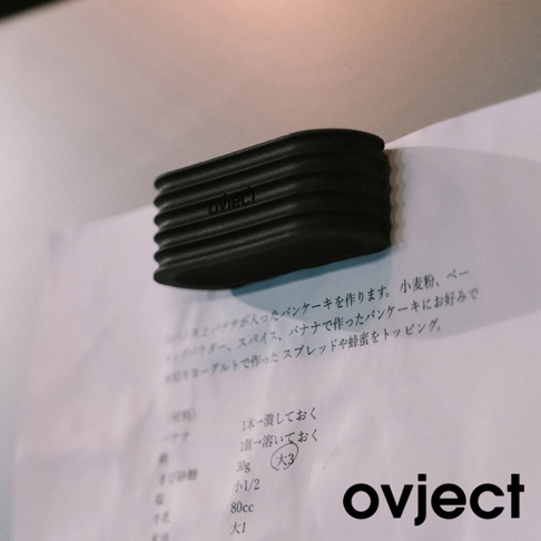 黑色帶有紋路的圓形鍋蓋，上面印有 ovject 字樣，部分蓋在印有日文食譜的紙張上。