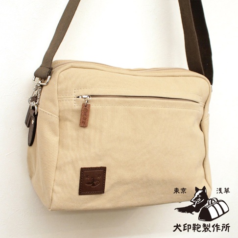 米色帆布斜背包，正面有拉鍊口袋，並飾有皮革標籤和印花圖案。