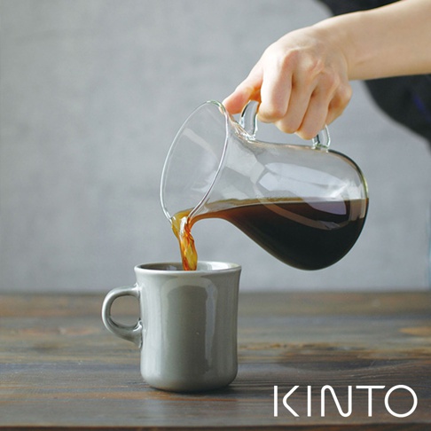 玻璃咖啡分享壺傾倒著深褐色咖啡液，注入一個灰色陶瓷馬克杯中。分享壺具有透明玻璃材質和彎曲的把手。