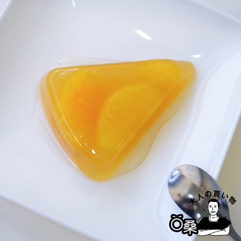 日本國產水果三角形果凍，呈現鮮豔的橘黃色，內含切片水果，果凍外層包裹透明的凝膠，置於白色盤中，旁有一金屬湯匙。