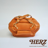 職人手作皮革輕旅行後背包，採用質感棕色皮革，設有兩條可調節的皮革提把，搭配金色金屬扣環和鉚釘，整體造型簡約實用。