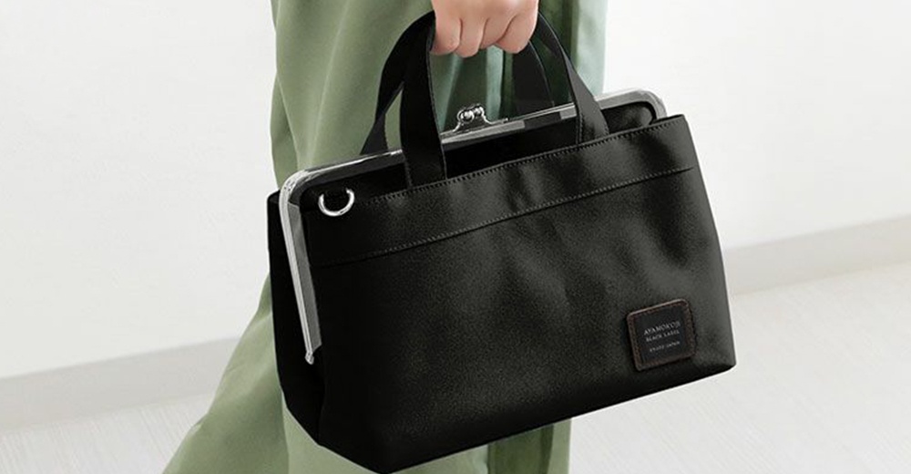 一位女士手提一個黑色的京都職人手作橫式口金手提包，包包上有品牌標籤 AYANOKOJI BLACK LABEL MADE IN JAPAN。