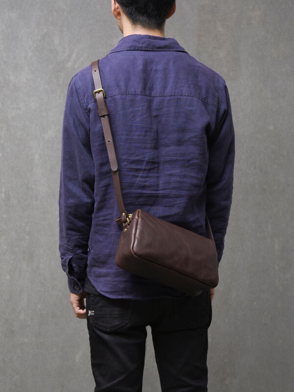 職人手作皮革Organ輕巧拉鍊單肩包 (G-80) M，深棕色荔枝紋皮革，帶有金色拉鍊與金屬配件，深棕色皮革肩帶可調節長度，單肩斜背於身著深藍色襯衫的模特兒身上。