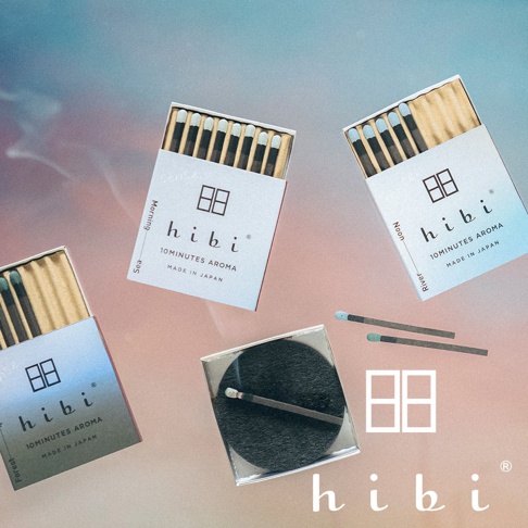 三盒標註著 hibi 品牌的日式香氛火柴禮盒散落在帶有漸層色彩的背景上，每盒標示著如晨、午、海、河、森林等情景主題，禮盒旁還放置了兩支散開的火柴，中央則有一個黑色圓形底座與一支已點燃或使用過的火柴，整體呈現簡約質感與日系文青風格。