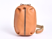 職人手作皮革立體運動斜背包，呈現自然淺棕色調。包身以光滑皮革製成，線條圓潤飽滿，中央有一條銀色拉鍊，搭配米色織帶和皮革拼接的斜背帶，整體風格簡約實用。