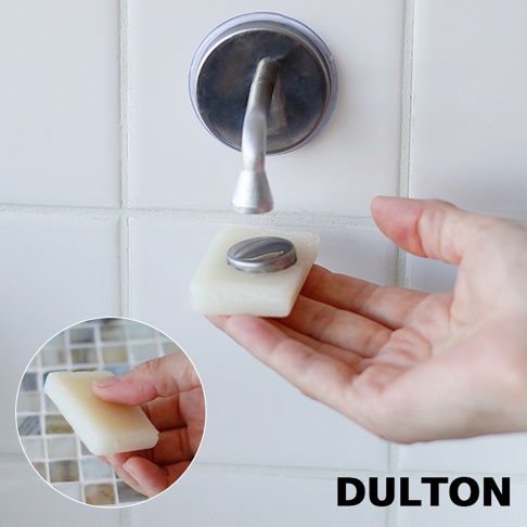 一塊白色方形肥皂，側面嵌有銀色金屬磁鐵，被一隻手托起，靠近牆上一個帶有銀色金屬鉤的圓形吸盤。