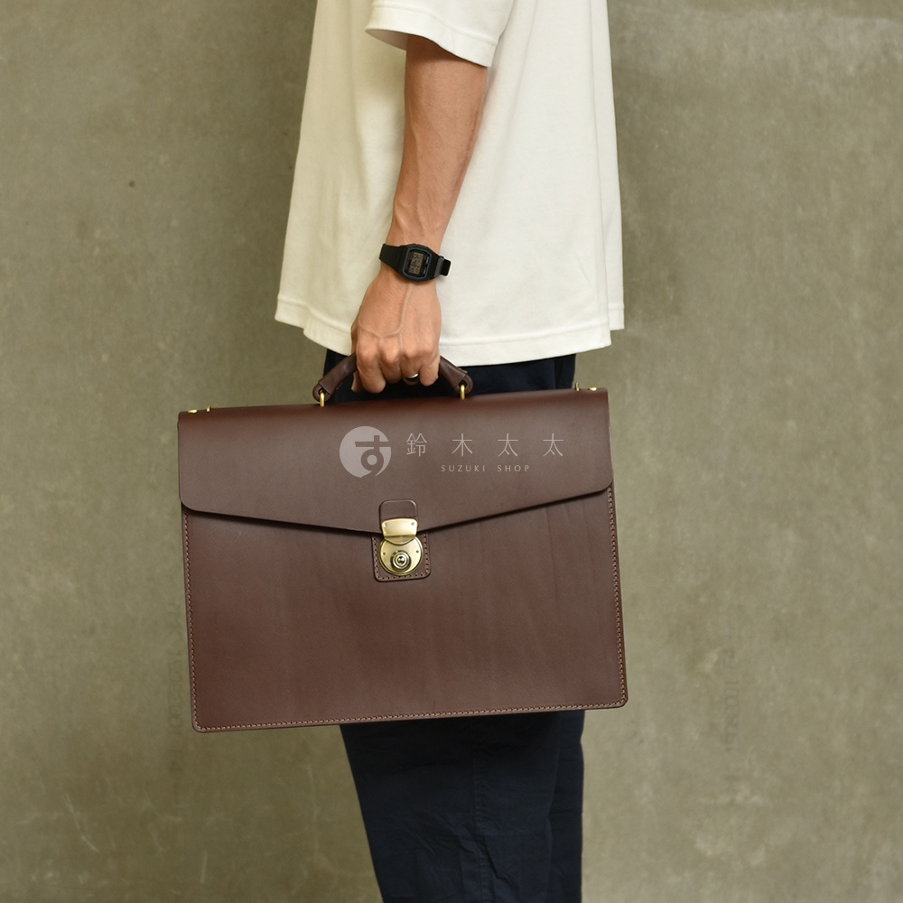 深棕色皮革公事包，配有頂部提把、金色鎖扣和縫線細節，由一人手持。