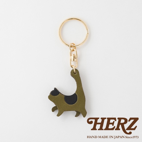 職人手作皮革貓咪造型鑰匙圈，橄欖綠色皮革，點綴兩塊黑色斑紋，搭配金色金屬掛環與勾扣，展現簡約可愛的貓咪姿態。