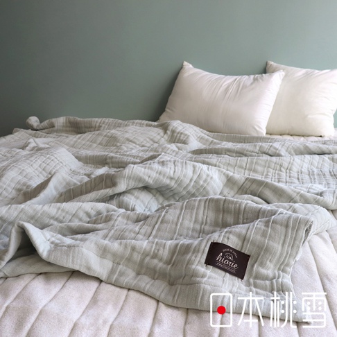 淺灰色八層紗雙人透氣被，材質為純棉，呈現褶皺紋理，帶有深棕色品牌標籤，放置於雙人床上，背景為淺綠色牆面，搭配兩個白色枕頭。