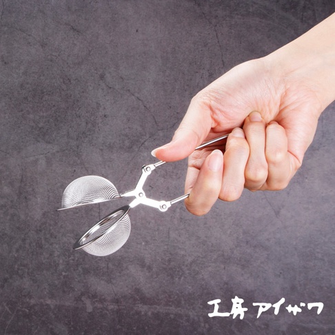 一只手拿著一個銀色不鏽鋼泡茶器，該泡茶器由兩個網狀球體組成，由鉗子狀的手柄連接。