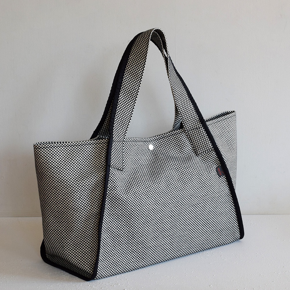 三河木綿刺子織可變形大容量托特包，以黑白格紋圖案織成，配有黑色滾邊和提把，包面中央有一個銀色按扣。