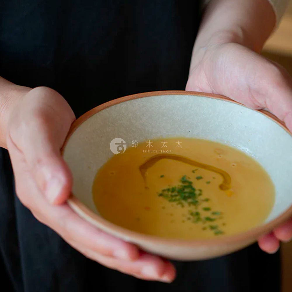 雙手捧著一個淺色陶瓷沙拉碗，碗內盛有金黃色濃湯，表面淋有橄欖油並撒有蔥花。碗的外緣呈陶土原色。
