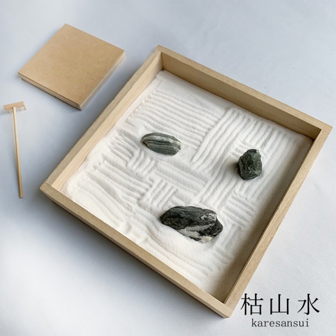 日式枯山水模型，木質淺色方型框，內含細白砂，砂上耙出紋路，擺放三塊深綠色帶有灰白色紋理的石頭。旁邊有木質底座和一把迷你竹耙。