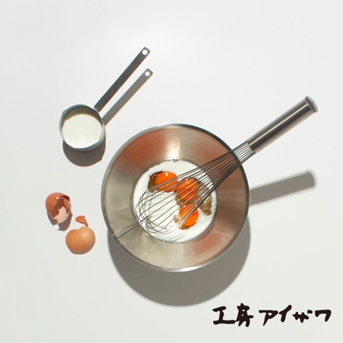 不鏽鋼雙口長柄計量杯，裝有液體，置於白色背景上，旁有打蛋盆、打蛋器、蛋和蛋殼。