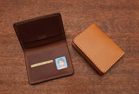 職人手作皮革雙折證件收納夾，一個為深棕色皮革，內有證件照與日文文字；另一個為淺棕色皮革，閉合狀態，兩者均以手工縫線製作。