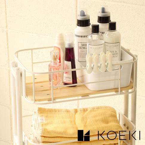 白色三層置物收納推車，上方層架擺放著粉色香水瓶、紫色瓶裝噴霧、白色與透明瓶身的清潔用品，以及白色夾子。下方層架則堆疊著幾條黃色毛巾。