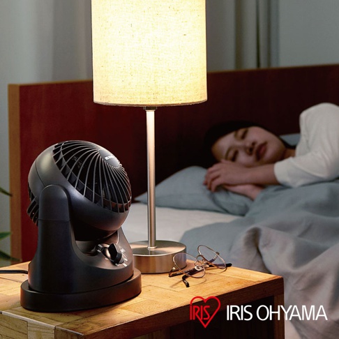 黑色迷你型空氣循環扇，帶有可調節的旋鈕，放置在床頭櫃上，旁邊有一盞檯燈和一副眼鏡，背景是睡覺的女性。