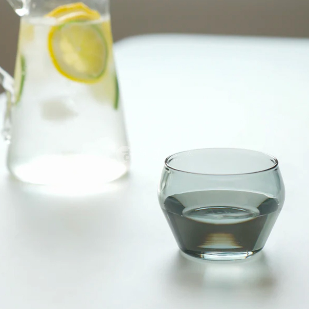 一個無色玻璃杯，杯身呈現圓潤的復古造型，裝有深灰色液體。背景是一個玻璃水壺，裡面裝有水和檸檬片。