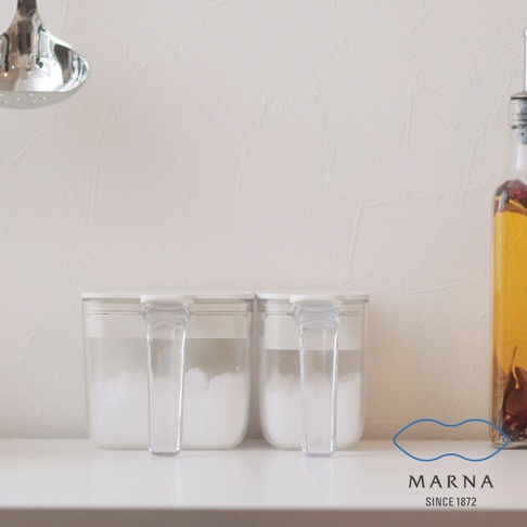 兩個極簡風格的白色透明密封調味料盒並排放在白色桌面上，容器呈現長方形，配有側邊把手，盒子下方裝有白色粉末狀調味料。背景為純白牆面，左上角懸掛一支金屬湯勺，右側則放有一瓶裝著辛香料的油瓶。右下方印有 MARNA SINCE 1872 的品牌標誌。