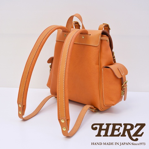 職人手作復古風皮革旅行後背包，呈現天然棕褐色皮革質感，配有雙肩帶、翻蓋和側口袋，以黃色縫線點綴，整體設計簡約耐用。