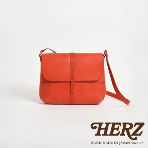 日本 HERZ 品牌橘紅色皮革細扣帶馬鞍包，搭配手工縫製細節，包蓋上有一個長方形小扣帶，可調整包包開合。