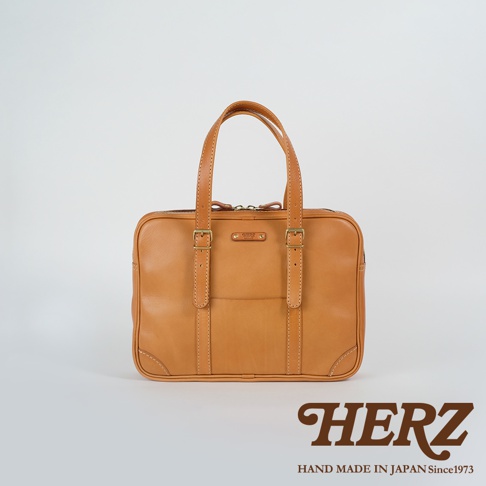 淺棕色皮革製成的職人手作公事包，可肩背或手提，配有頂部提把、兩個可調節的皮革束帶及金色扣環，正面有一個開放式口袋。
