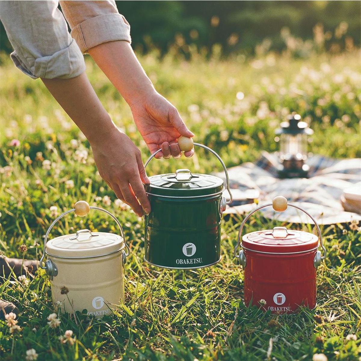 綠色、米色和紅色的 OBAKETSU 蚊香桶放在草地上，其中一個綠色桶被一只手拿起。