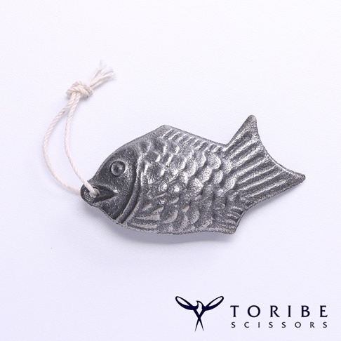 銀灰色金屬製成的小鯛魚造型，帶有一條米白色細繩，繩子末端打結。鯛魚表面刻有魚鱗紋路，尾部呈扇形。