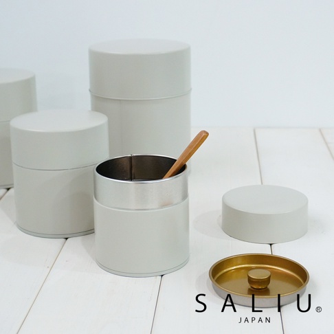 淺灰色簡約錫製茶葉罐，其中一個罐子內有一支木質勺子。周圍散落著兩個同款茶葉罐和一個金色內襯的淺灰色罐蓋。