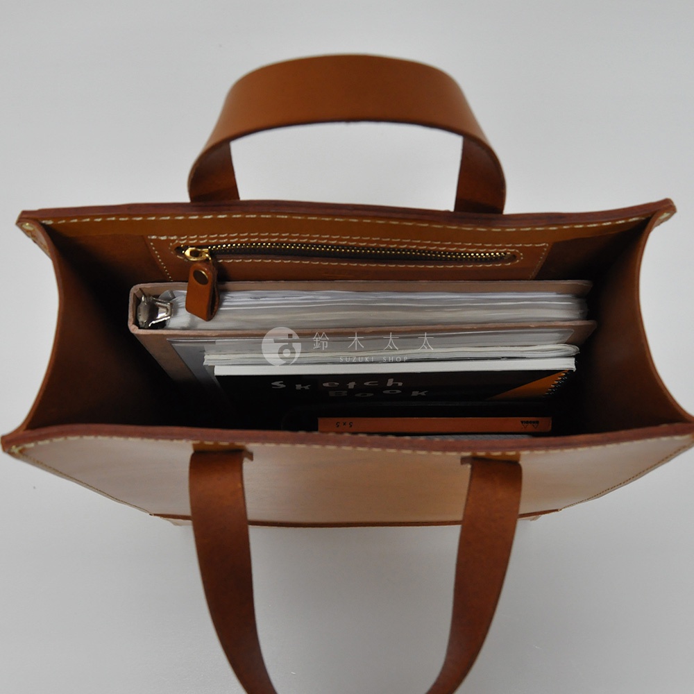 職人手作皮革直式托特包，棕色皮革材質，包內裝有筆記本和素描本，包口設有拉鍊，附有兩條皮革提把。