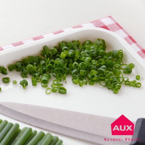 白色迷你砧板上擺放著切碎的蔥段，旁邊是銀色刀刃的菜刀，背景是紅白色格紋布。