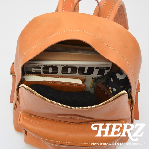 職人手作皮革休閒風後背包，展示內部空間，可見雜誌、筆記本、黑色毛衣、錢包等物品，背包為鞣製棕褐色皮革，搭配縫線與黃銅色拉鍊。