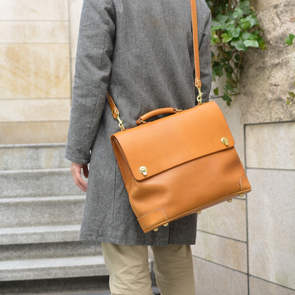 淺棕色皮革製成的波士頓包，附有可拆卸肩帶，包包上配有兩個金色金屬旋轉鎖扣和提把，邊角有金色金屬保護釘。