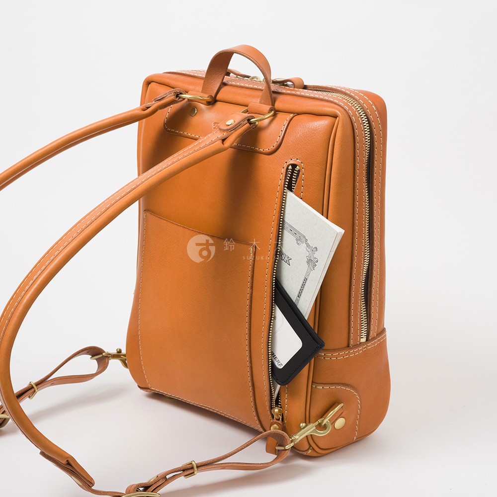 職人手作的棕褐色皮革軟皮商務後背包，細緻縫線，前方有拉鍊口袋，內含一本白色筆記本和黑色卡夾。