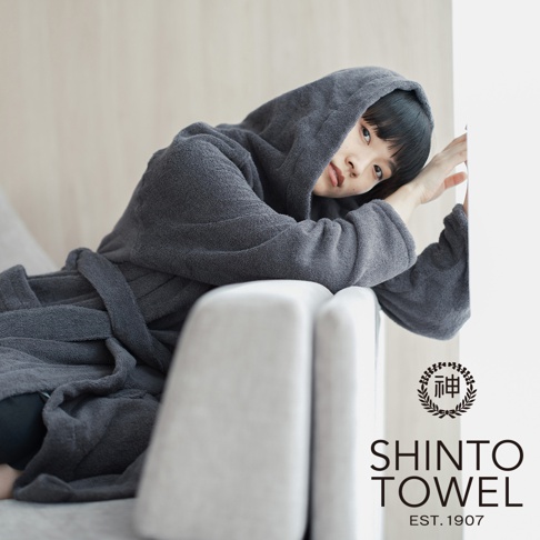 一位年輕女性穿著一件深灰色連帽蓬鬆浴袍，斜倚在白色沙發上，浴袍材質為有機棉，觸感柔軟。